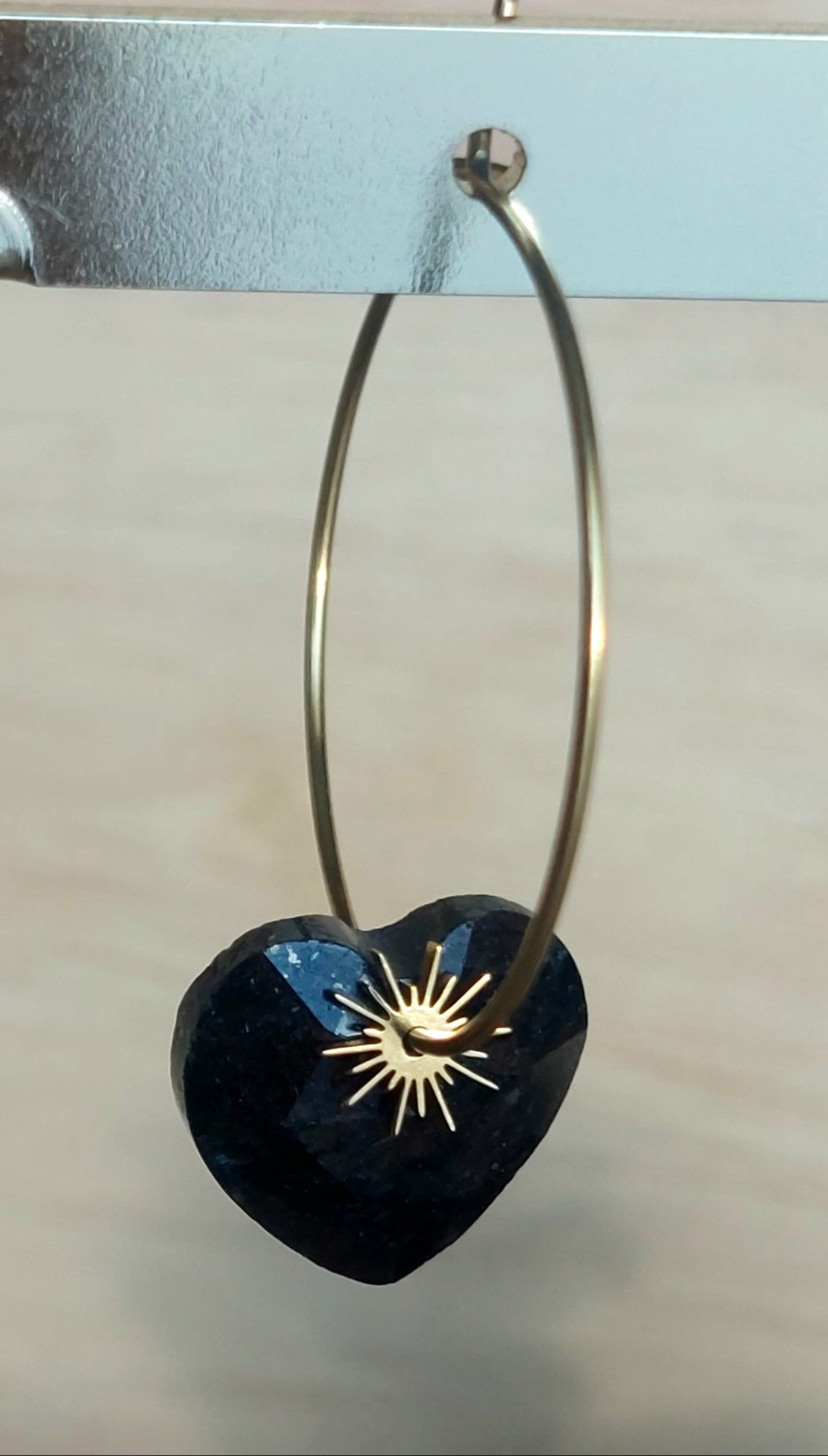 Zoom sur des créoles acier inoxydable ornées d’un cœur en pierre naturelle  tourmaline noire et d’un petit soleil brillant.