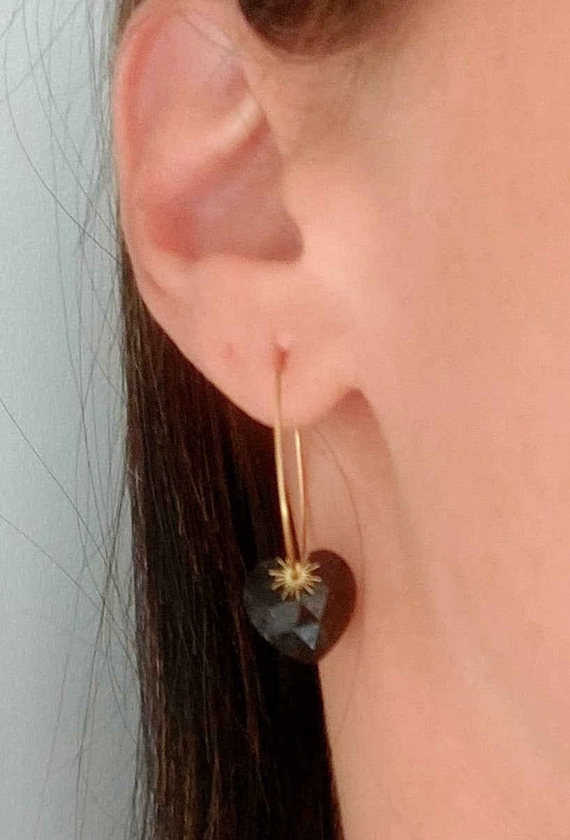 Boucles d’oreilles créoles en acier inoxydable portées, avec pendentif cœur tourmaline noir et soleil doré.