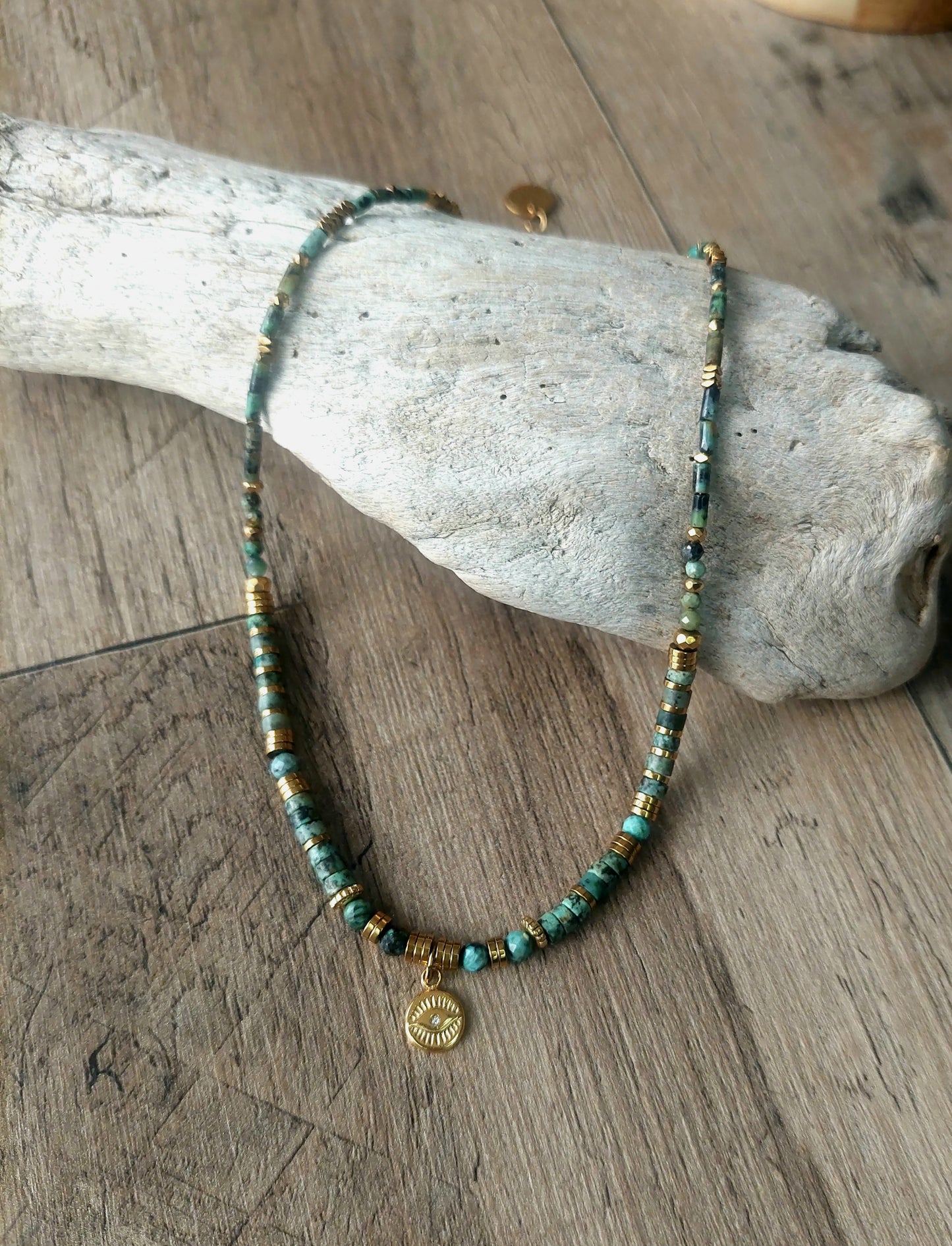 Collier bohème en perles heishi turquoises africaines avec pendentif œil protecteur, délicatement présenté sur bois flotté, symbole d’élégance naturelle et de savoir-faire fait main – modèle unique.