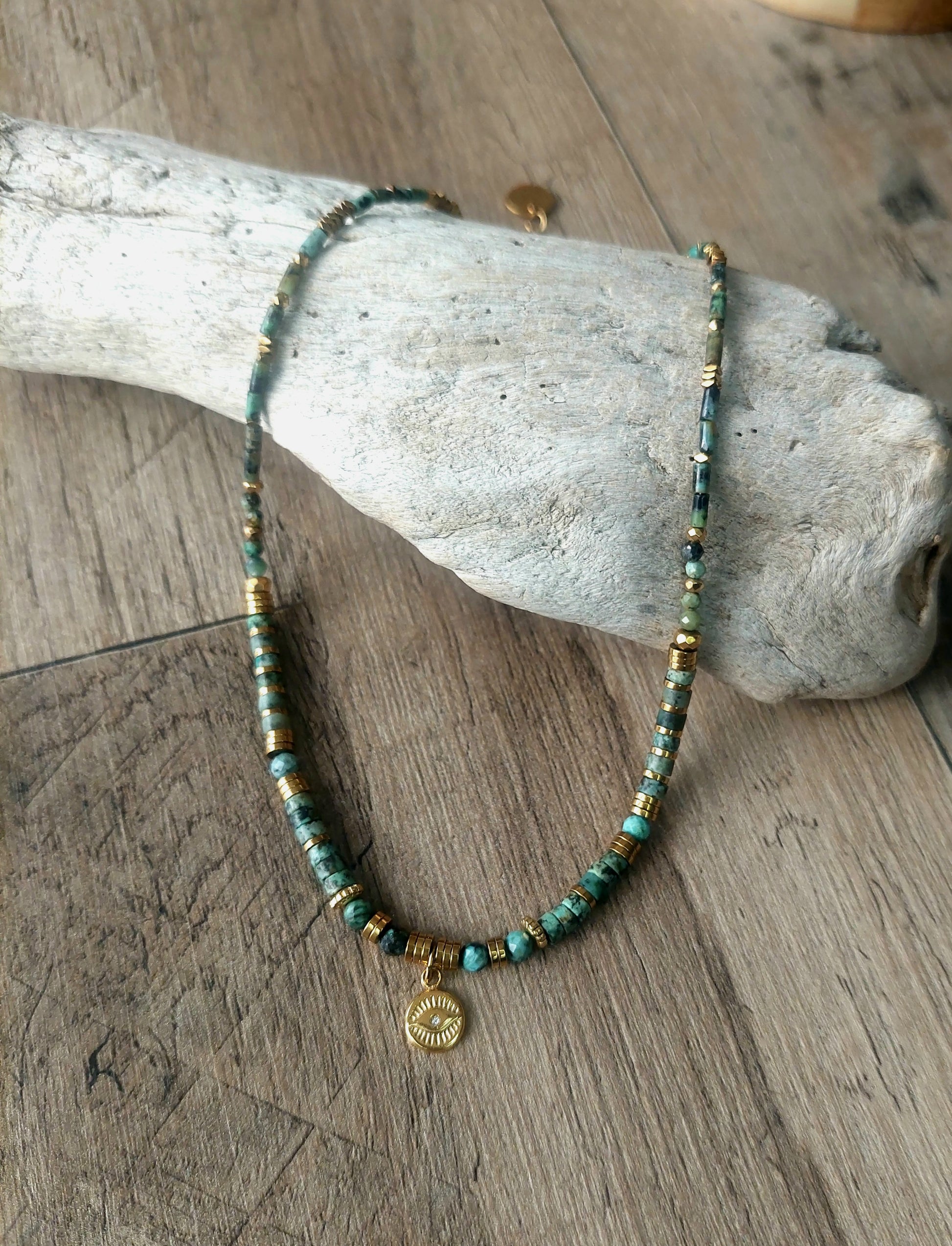Collier bohème en perles heishi turquoises africaines avec pendentif œil protecteur, délicatement présenté sur bois flotté, symbole d’élégance naturelle et de savoir-faire fait main – modèle unique.