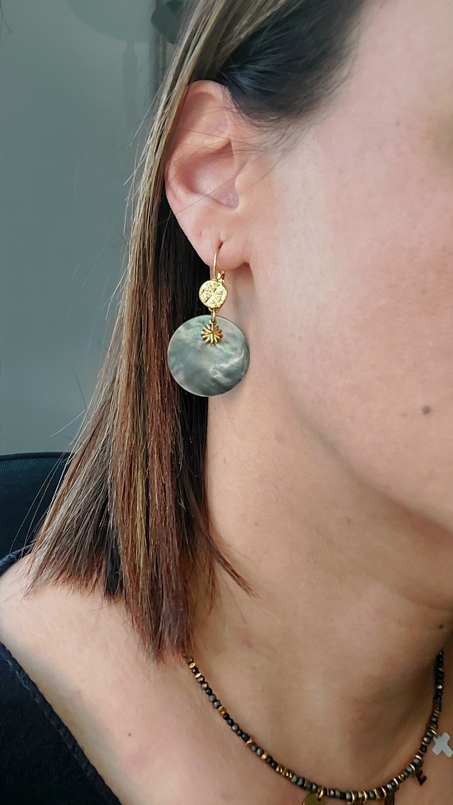 Femme portant des boucles d’oreilles dormeuses chics et bohèmes en acier inoxydable avec nacre naturelle et pendentif soleil fait main